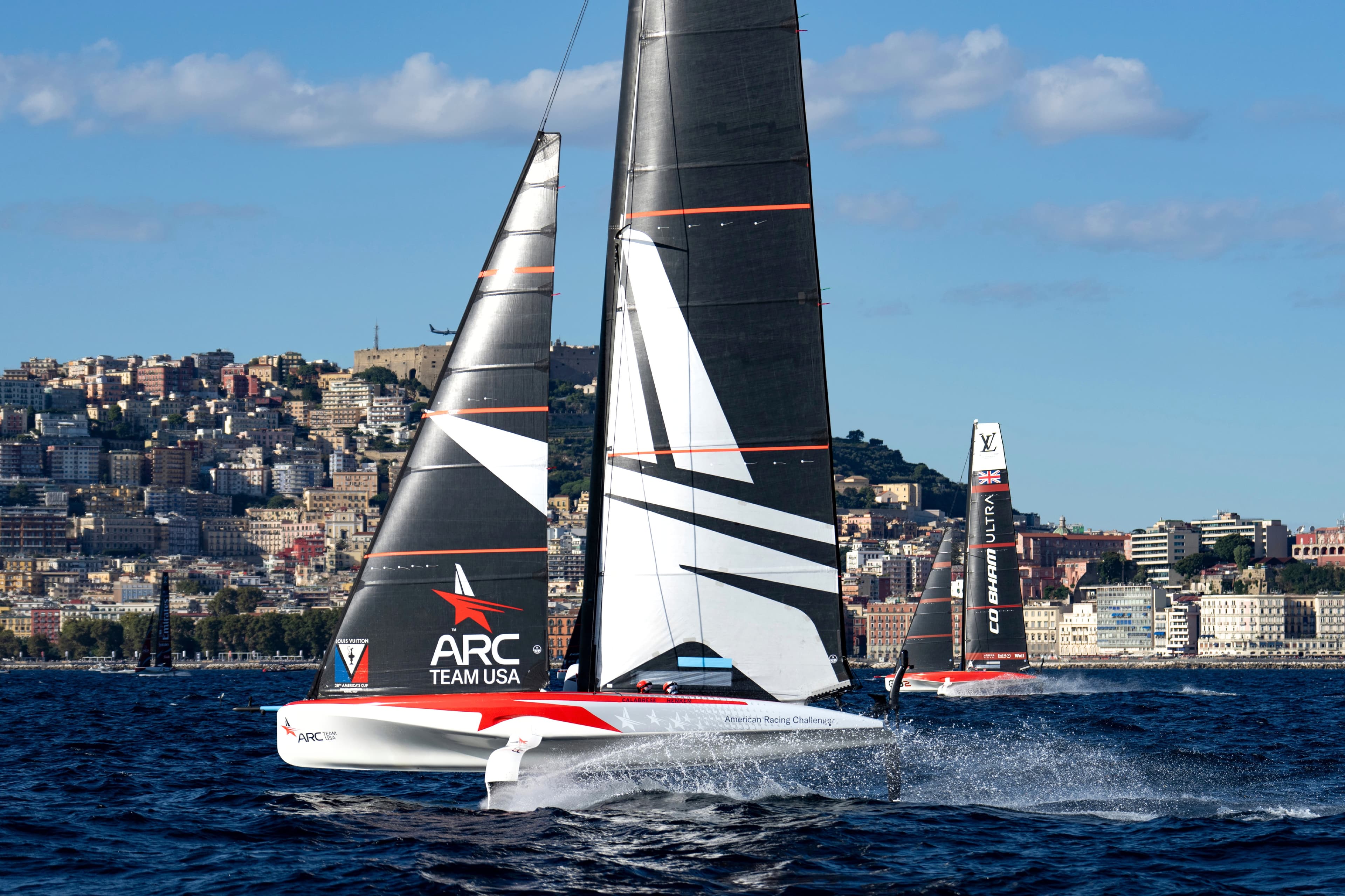 ARC Team USA's AC40 foiling on the Bay of Naples during the Louis Vuitton 38th America's Cup Preliminary Regatta, with the Naples waterfront behind.